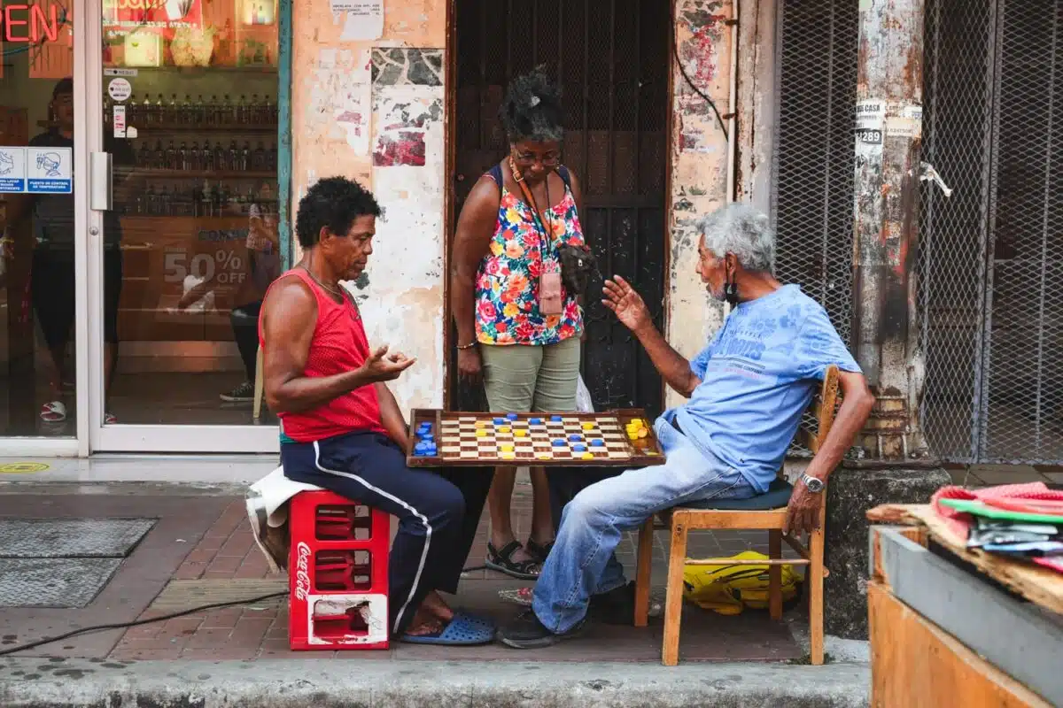 A woman interrupting two men playing chess with bottle caps in Santa Ana neighborhood