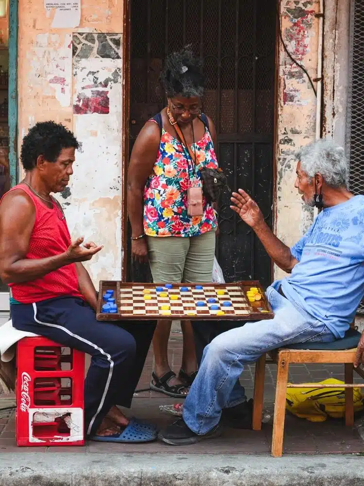 A woman interrupting two men playing chess with bottle caps in Santa Ana neighborhood